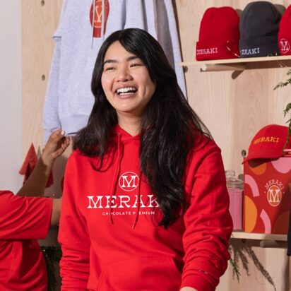 Smiling staff wear red coloured merch for Meraki (chocolate premium) as they stock a store's shelves.