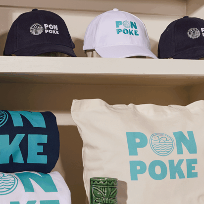 Custom branded hats and t-shirts in blue, white, and black with Pon Poke logo, including a matching pillow – themed merchandise display.
