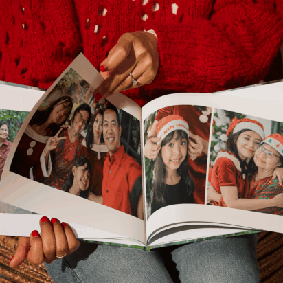 Woman holding a photo book with family Christmas pictures, surrounded by presents and a Christmas tree — personalized Christmas gifts idea.