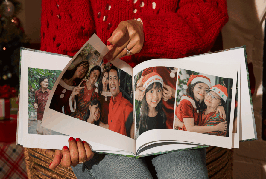 Woman holding a photo book with family Christmas pictures, surrounded by presents and a Christmas tree — personalized Christmas gifts idea.