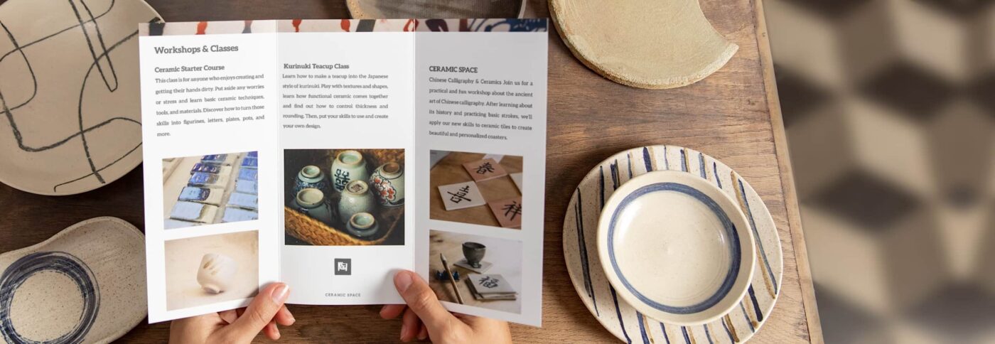 Hands holding a tri-fold brochure on ceramic workshops, surrounded by handmade pottery on a wooden table – example of brochure folds in real use.