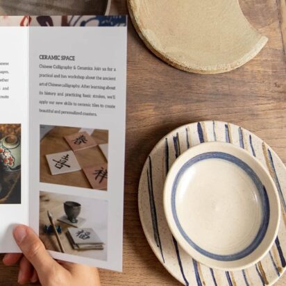 Hands holding a tri-fold brochure on ceramic workshops, surrounded by handmade pottery on a wooden table – example of brochure folds in real use.