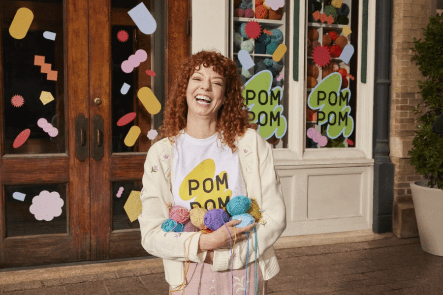Store owner smiling outside a yarn shop with branded window decals and a matching logo T-shirt