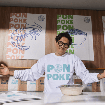 Smiling small business owner behind a branded poke counter, wearing a matching sweatshirt and surrounded by consistent packaging and signage