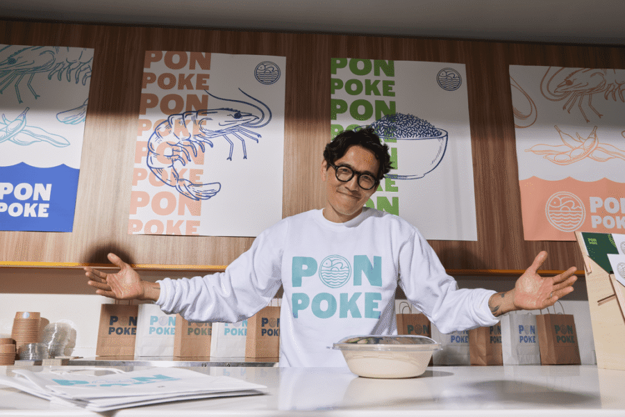 Smiling small business owner behind a branded poke counter, wearing a matching sweatshirt and surrounded by consistent packaging and signage