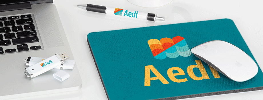 Branded merchandise on a white desk including a mug, USB stick, pen, and mousepad printed with a colorful logo