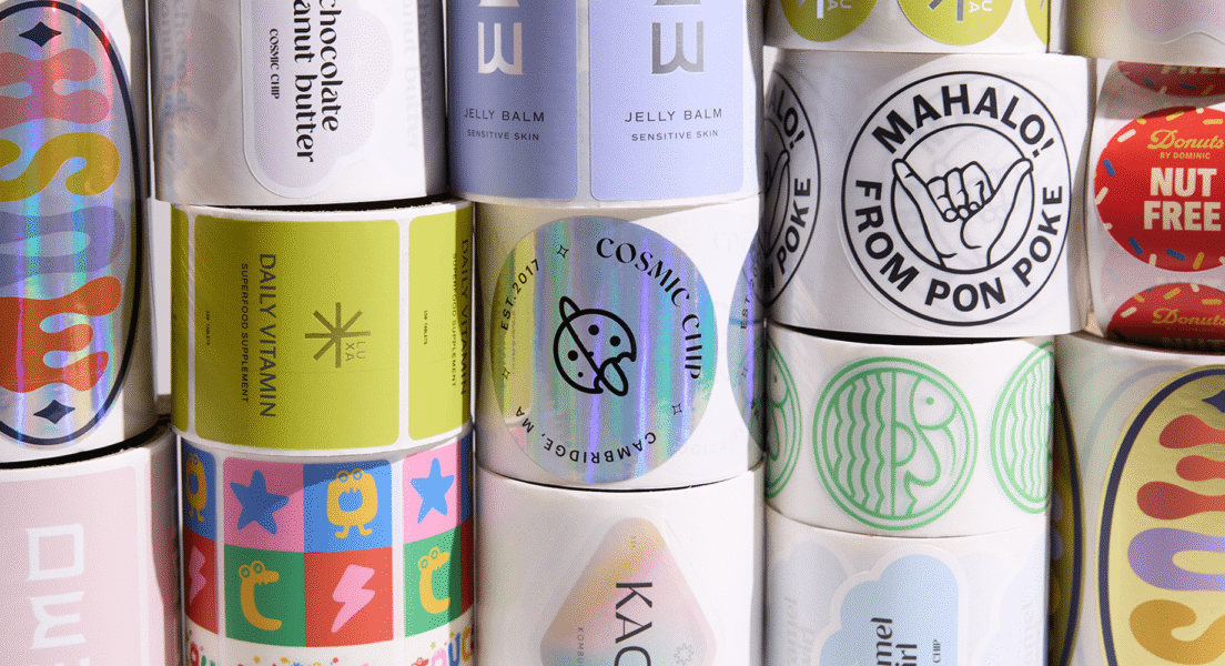Multiple rolls of colorful custom stickers and logo labels stacked in a grid pattern, showing a variety of brand logos and designs.