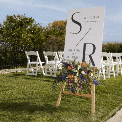 Wedding welcome sign with colorful floral arrangement on a wooden easel, set on a lawn with white ceremony chairs.