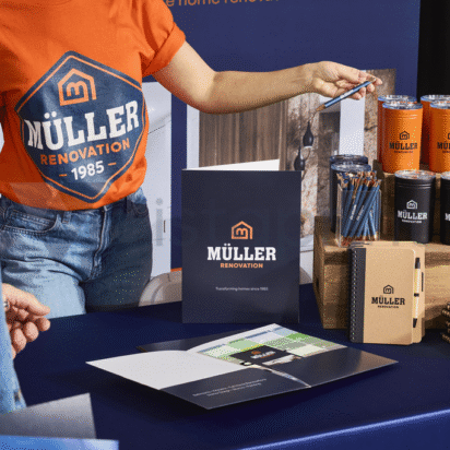 A branded collection of swag and merch from a Muller renovation company, notebook, pencils, drink containers, baseball hats and more arranged in layers across on a surface.