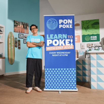 Man standing next to a blue retractable banner promoting a “Learn to Poke” class at Pon Poke, inside a modern and colorful restaurant space.
