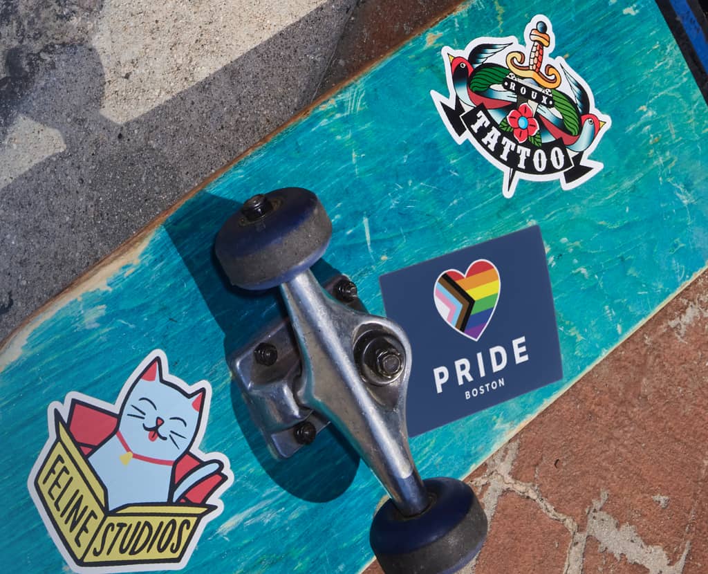Pride sticker on bottom of blue skateboard with other colorful stickers includes a heart-shaped pride flag.