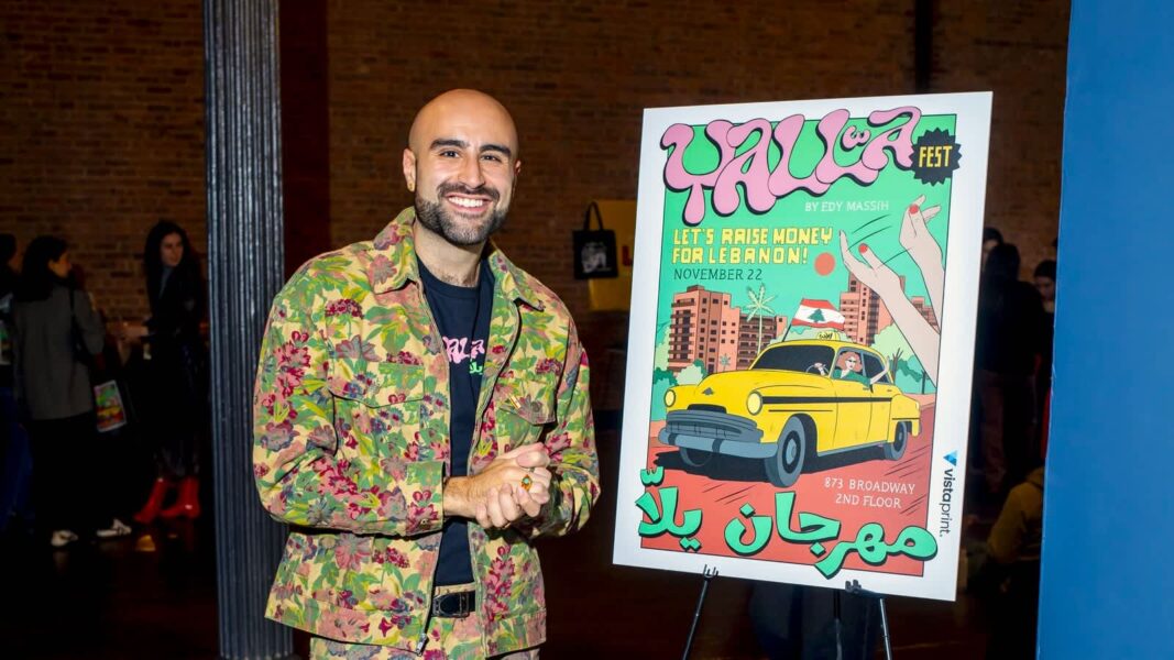 Man in colourful clothing stands smiling beside a large colourful poster advertising a community festival that he helps to organise - Yalla