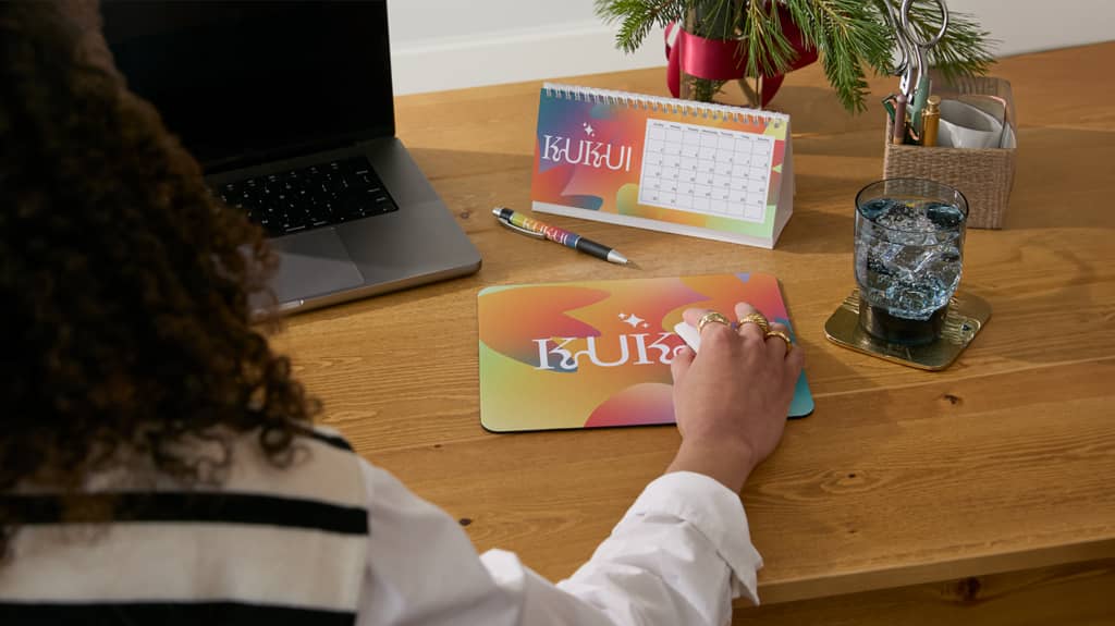 A woman using branded merch products at a desk: a mouse mat, pen and calendar