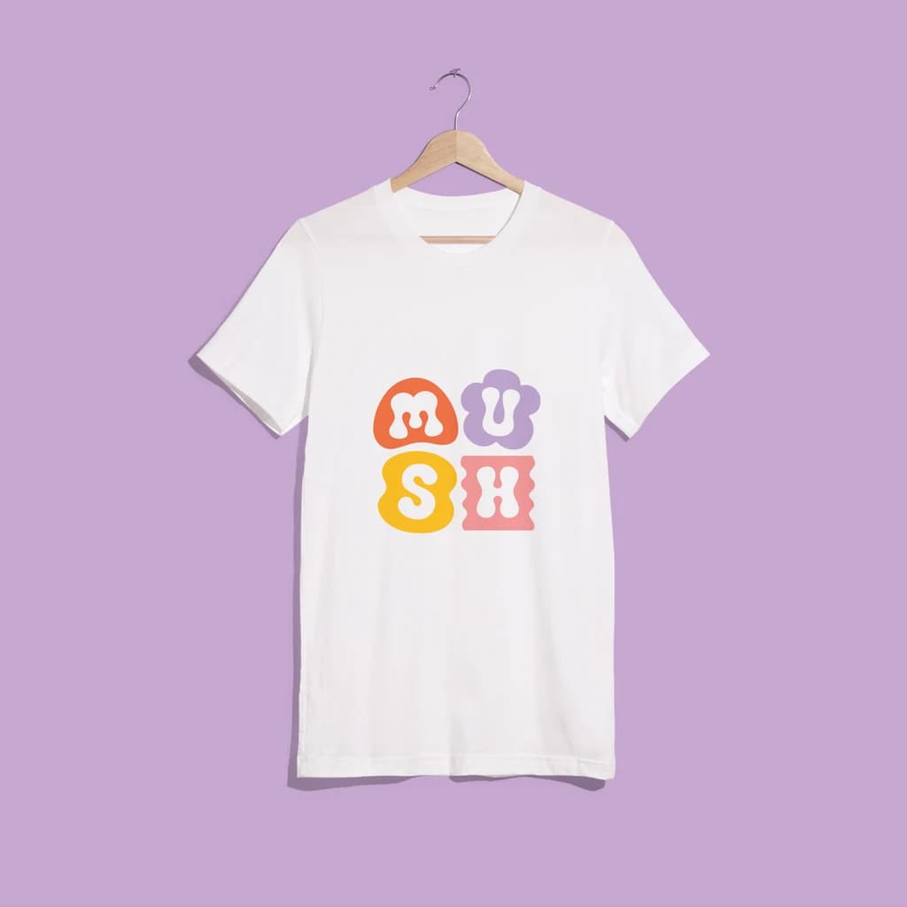 Branded T-shirt with colorful, square "MUSH" logo hangs against a lavender wall