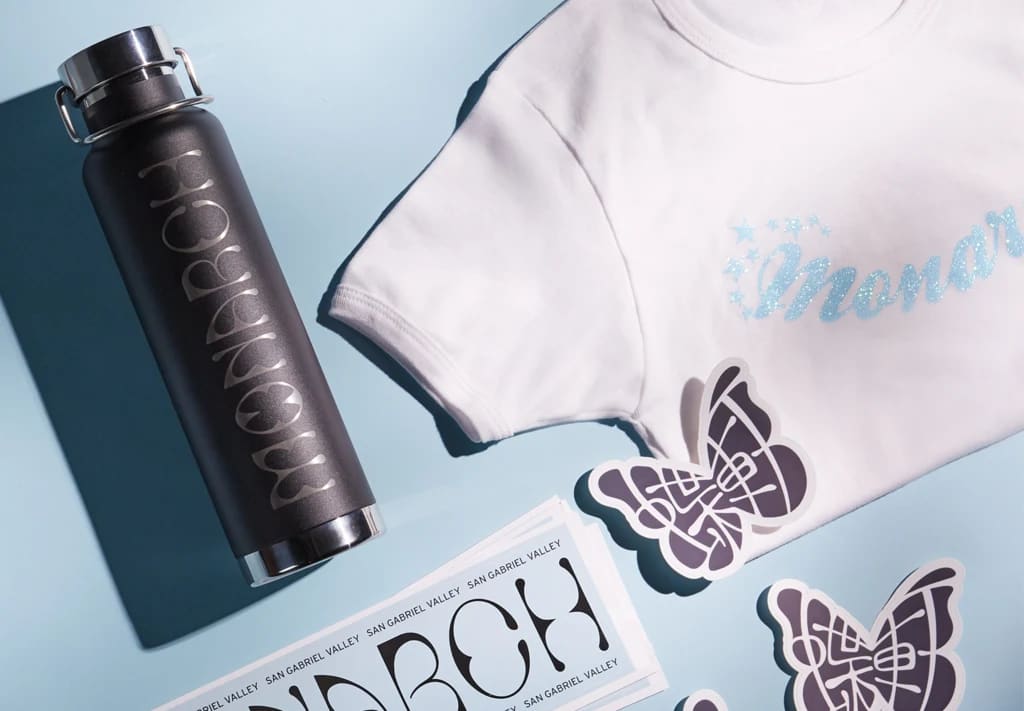 Promo items branded "Monarch" include coordinated water bottle, T-shirt and stickers setting on sky blue surface