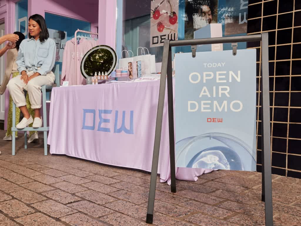Outdoor event marketing setup with a branded DEW table display and signage reading &ldquo;Open Air Demo,&rdquo; where staff engage attendees and showcase products in front of a storefront