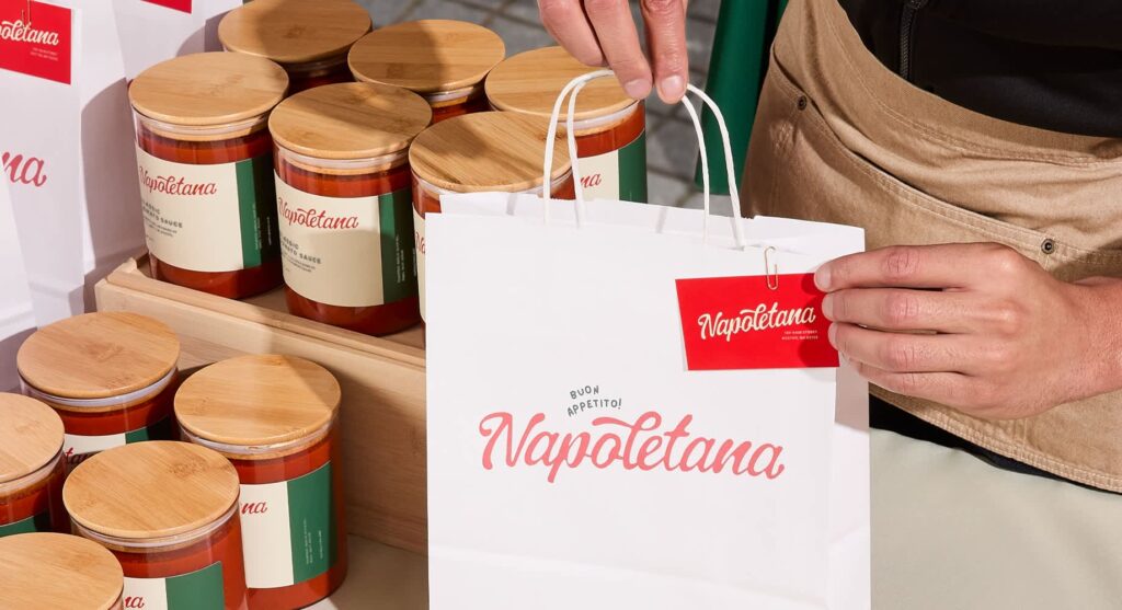 Close-up of a person placing a branded tag onto a white Napoletana paper shopping bag, with jars of tomato sauce displayed on wooden stands beside it