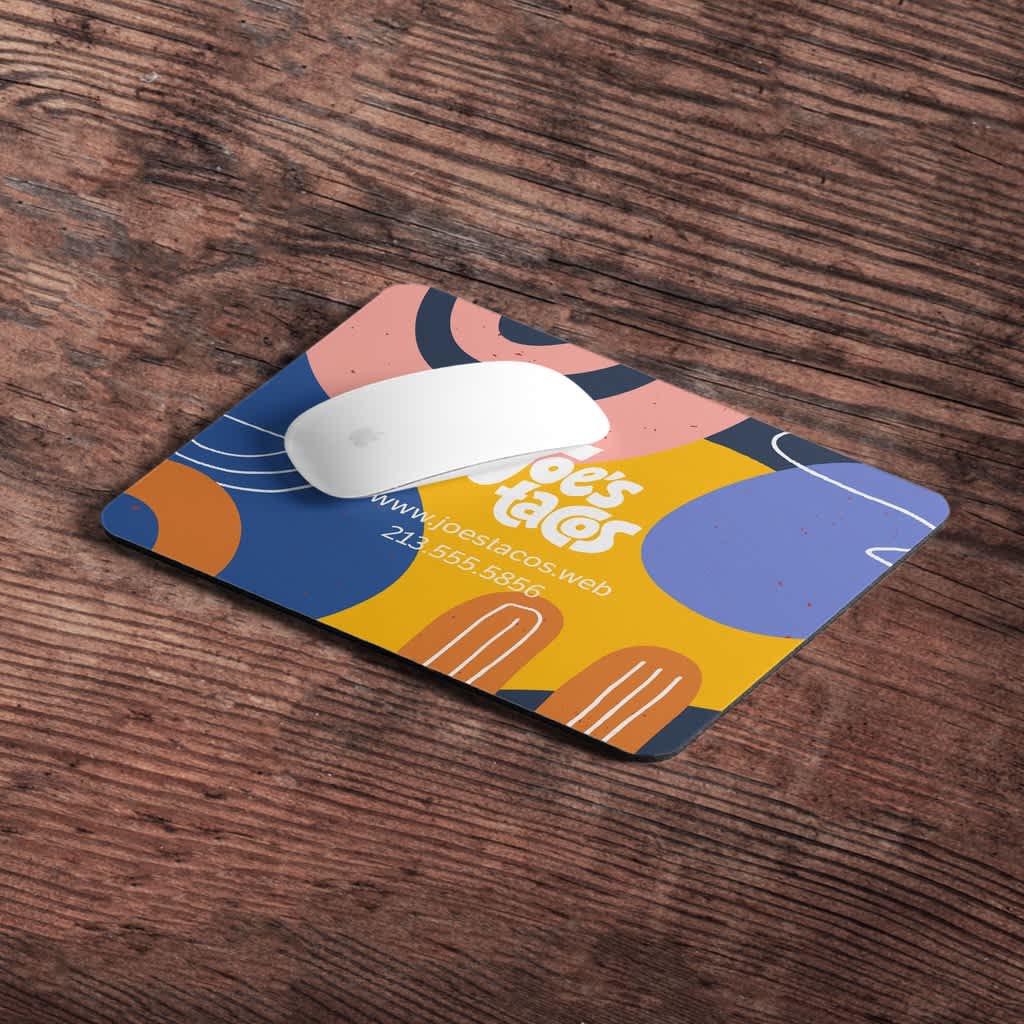 A white computer mouse on a colorful mousemat with branding for a taco company on a brown wooden table