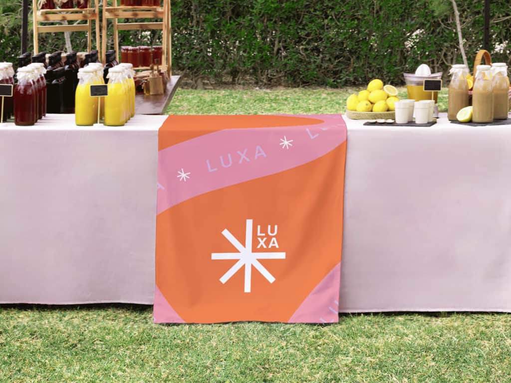 A pink table with an orange branded table cloth and bottles of juices at an outdoor event
