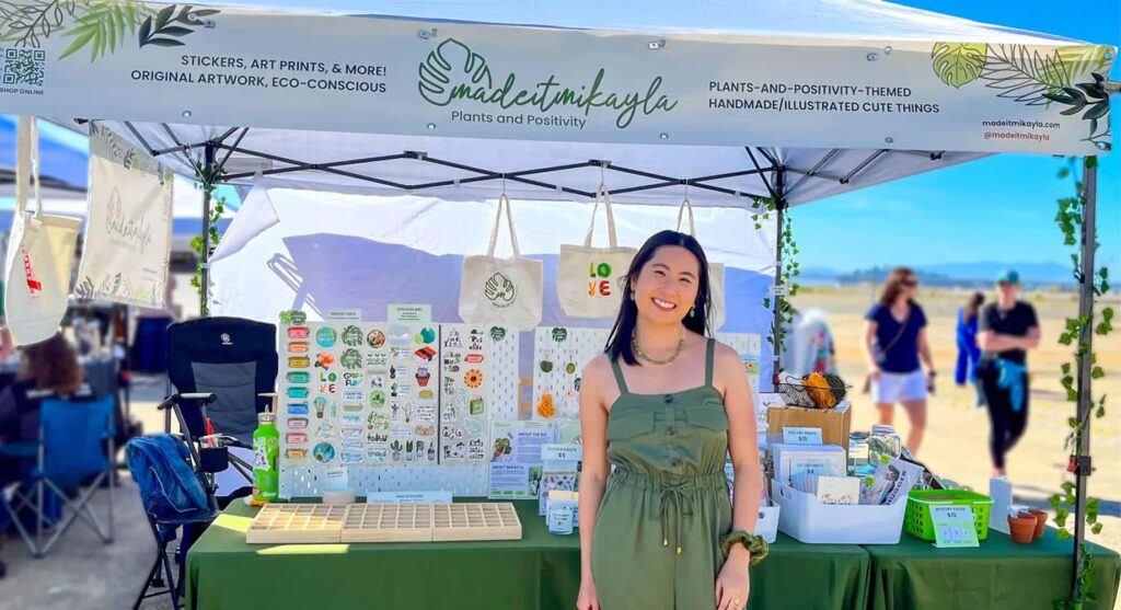 A woman standing in front of an outdoor market event marketing booth for Made It Mikayla