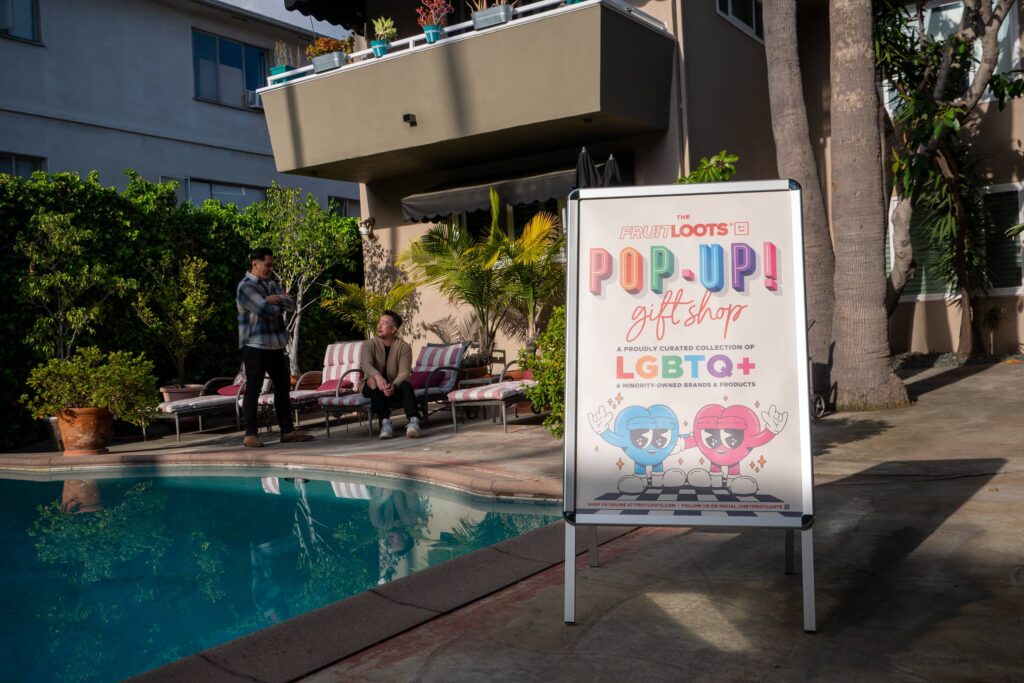 A colorful A-frame sign promoting a pop-up gift shop next to a swimming pool which two people are sat next to