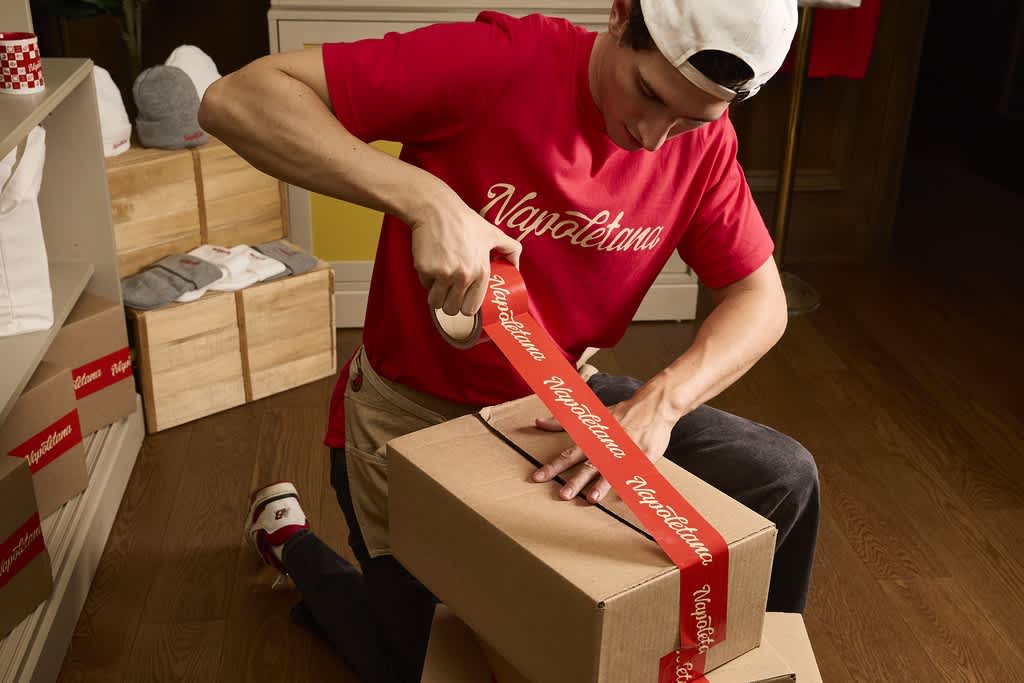 A person in a red branded t-shirt sealing cardboard box packaging with red tape.