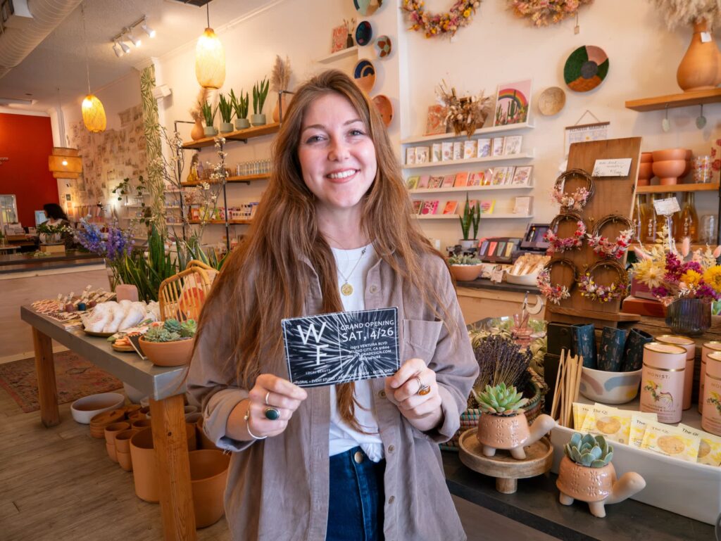 Small business owner in her shop looking at the camera and holding up a printed flyer for the grand opening