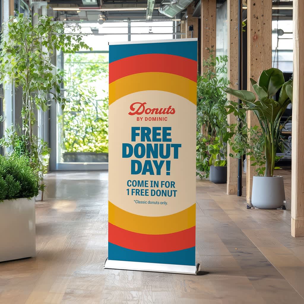 Retractable banner in a foyer surrounding by indoor plants with a simple blue, yellow and red design promoting a free donut day.