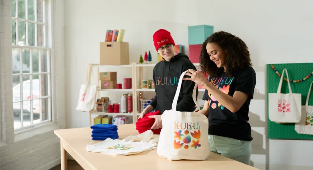 Two small business owners wearing branded merch and putting products into a tote bag.