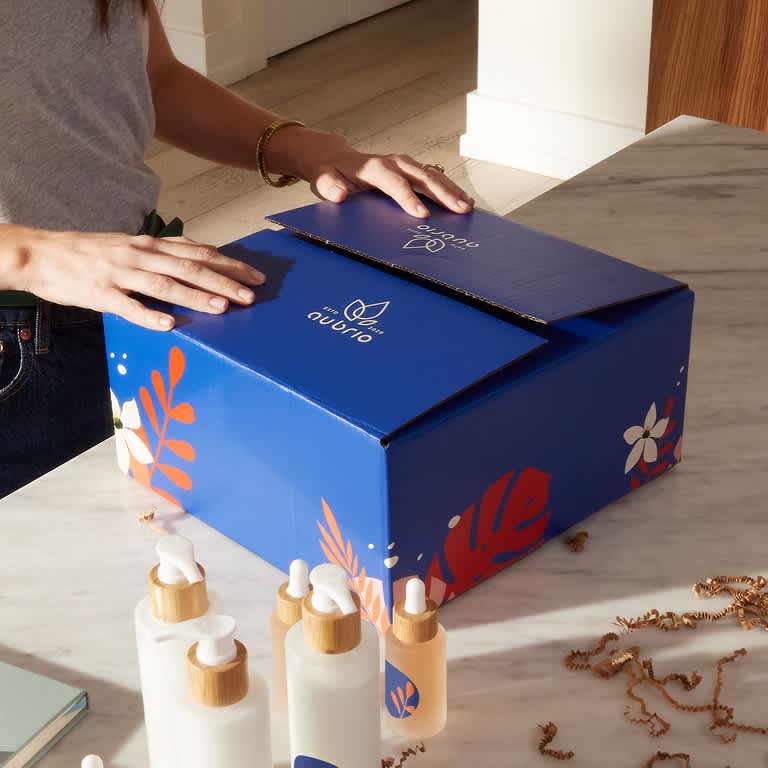 Woman opening branded shipping box with clean sans-serif typography and bold graphic packaging design for a skincare product set.