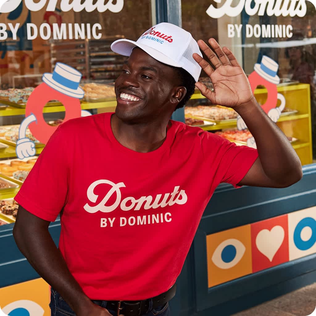 Donut shop employee wearing white embroidered trucker cap and red T-shirt with simple white “Donuts by Dominic” logo.