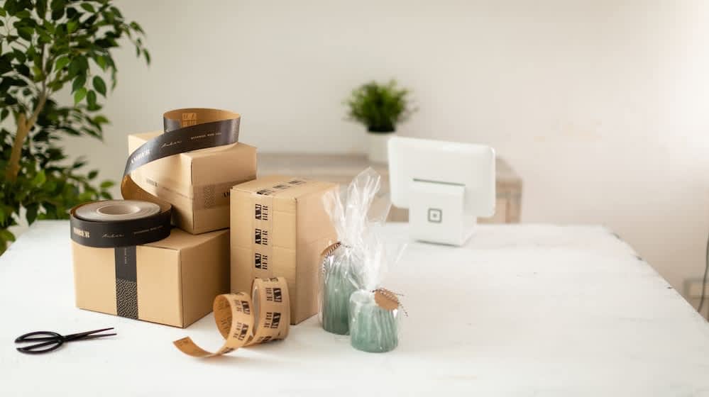 Two small candles in plastic wrap next to a brown box with branded packing tape.

