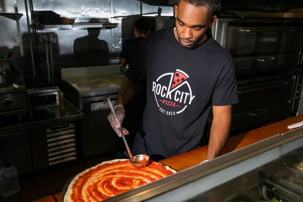 Chef applying tomato sauce to pizza base wearing black T-shirt with white and red pizza logo &ldquo;Rock City Pizza&rdquo;.