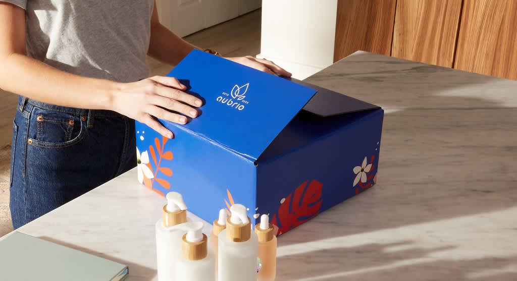 Hands closing a blue cardboard box package next to a collection of cosmetics bottles.