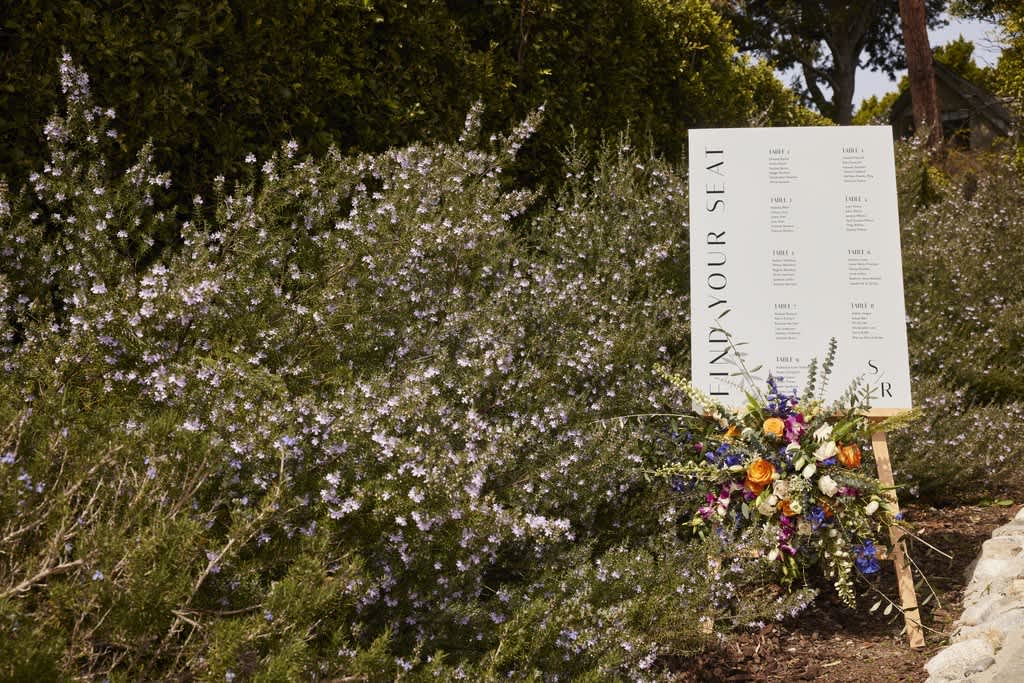 An outdoor wedding seating board surrounded by flowers and shrubbery.