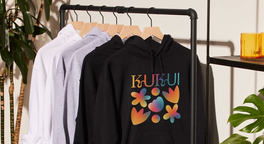 Selection of hoodies on a rack, the front one black with flower shape design and &ldquo;Kukui&rdquo; font all in rainbow palette.