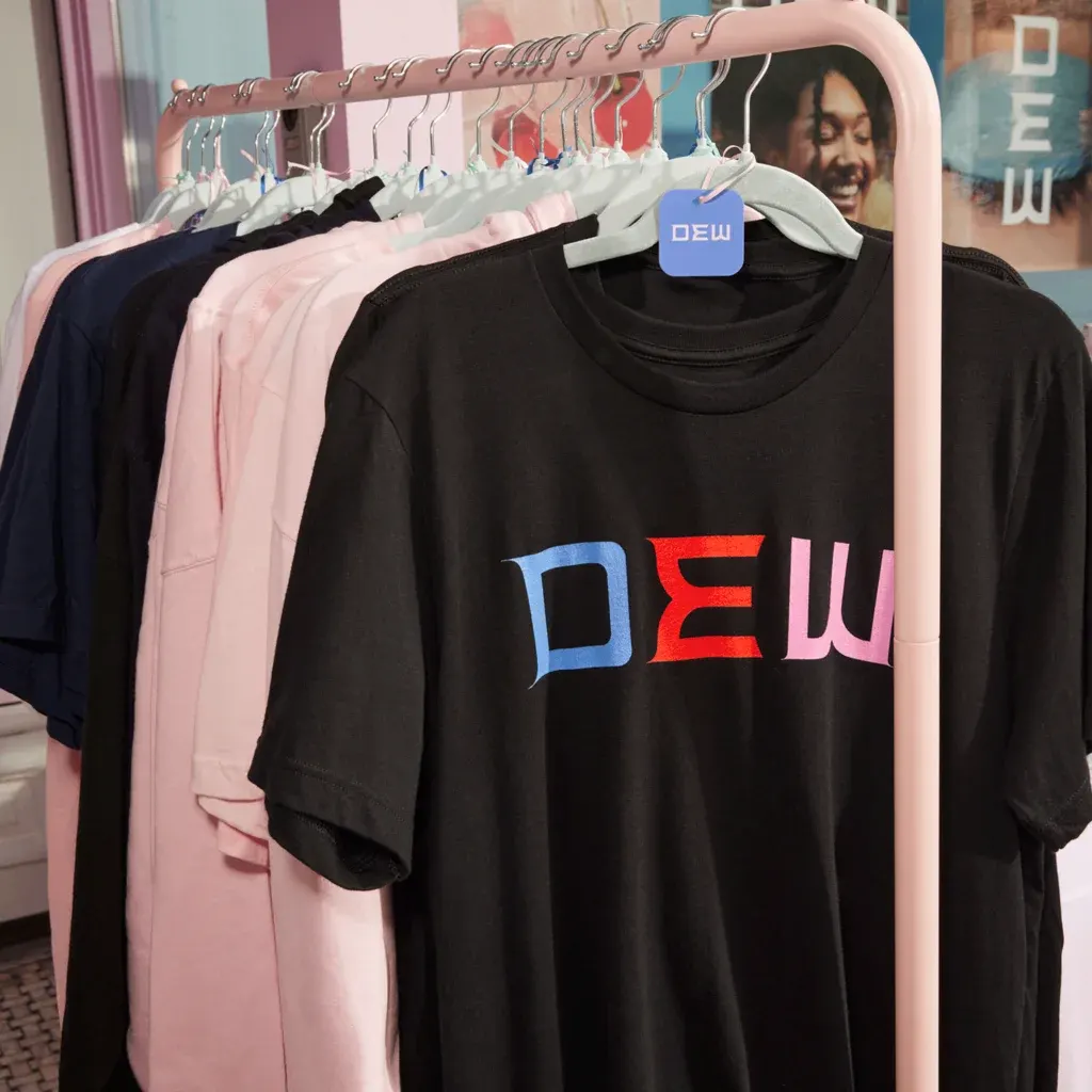 Selection of T-shirts and sweaters on pink clothes rack, the front being a black T-shirt with “DEW” font in blue, red and pink letters.