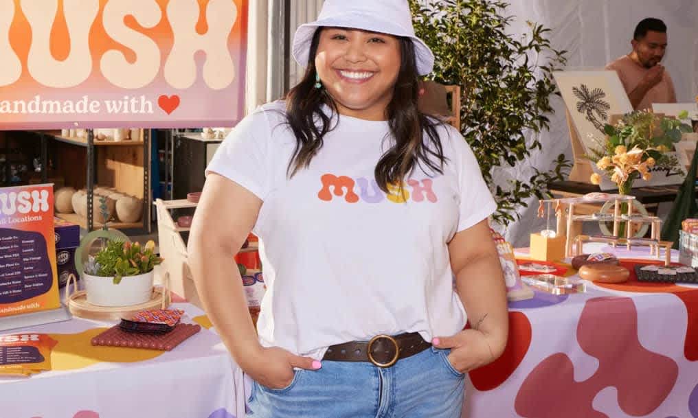 Small business owner smiling in front of event stall with various products and signage, wearing white T-shirt with “MUSH” logo in red, lilac, yellow and pink.