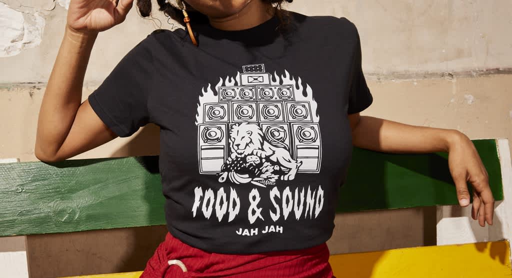 Small business owner wearing black branded T-shirt with pure white design depicting a large speaker sound system with a lion on a pile of fruit and “food and sound” underneath.