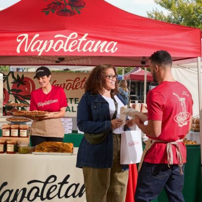 Staff wearing red branded apparel while serving customers at a busy outdoor market.