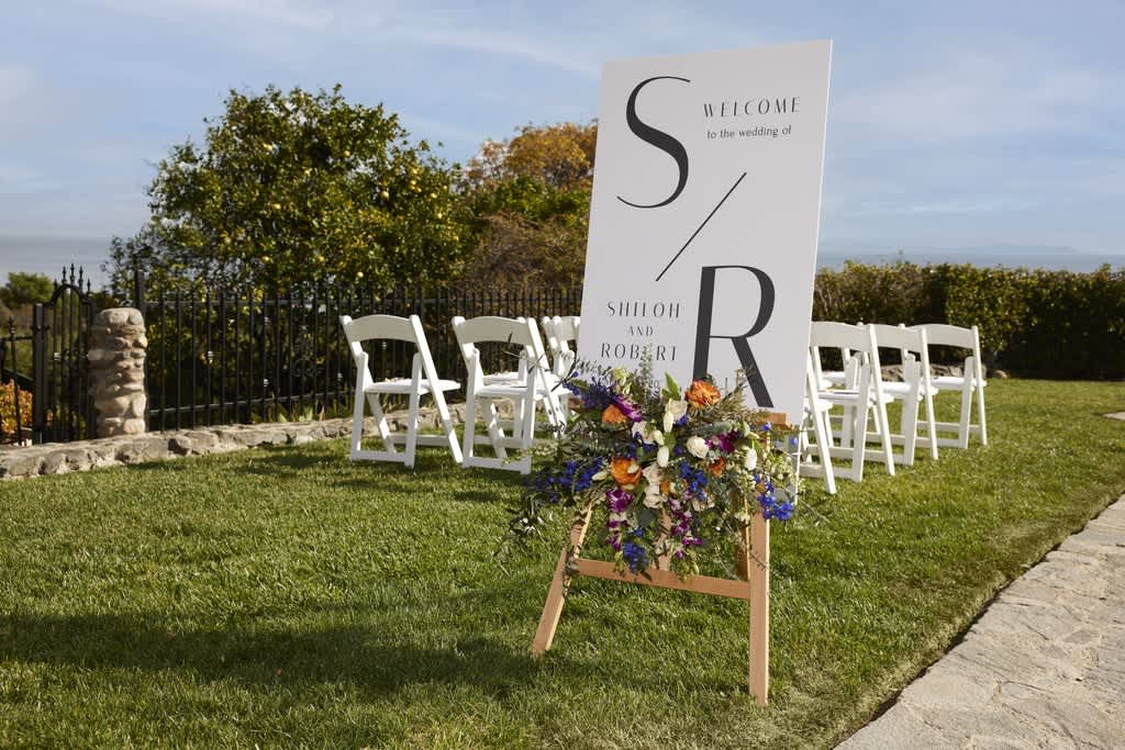 A wedding board with a bold S/R logo at an outdoor ceremony.