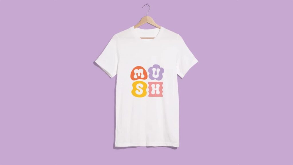 White T-shirt on lilac background with logo “MUSH” featuring organic shapes in red, lilac, yellow and pink for each letter.