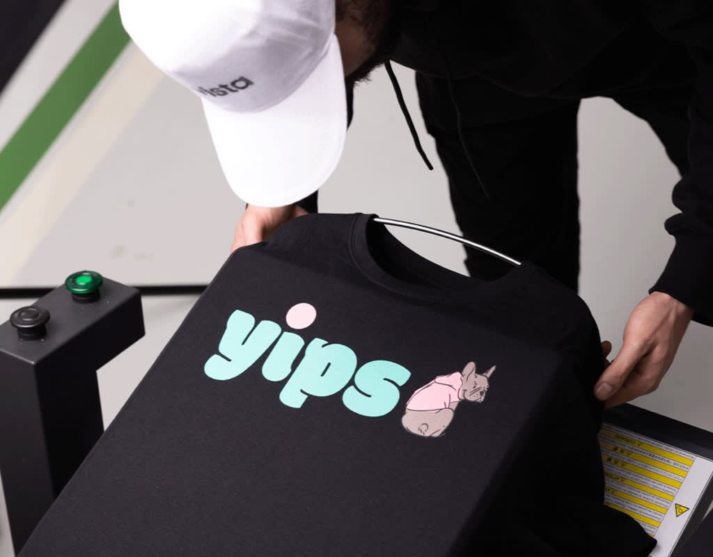 Man holding black T-shirt with dog graphic and light blue font &ldquo;YIPS&rdquo; over a printing press.