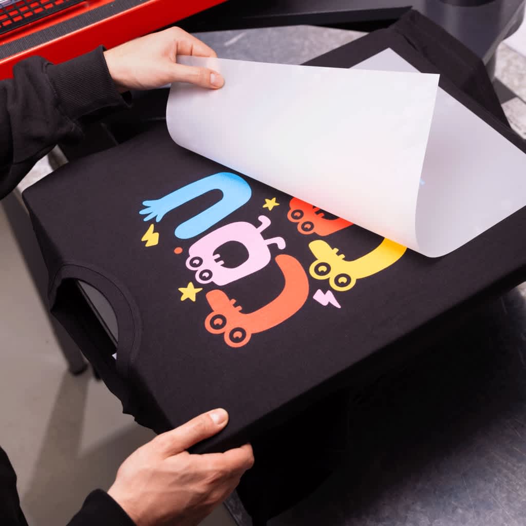 Someone removing transfer paper from a printing press making a black T-shirt with a neon shape graphic font &ldquo;COU.&rdquo;