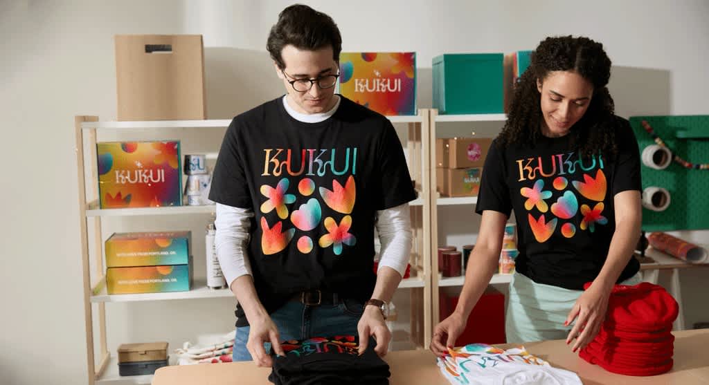Two small business owners folding stock in front of shelves wearing matching black T-shirts with colorful floral designs and font &ldquo;KUKUI.&rdquo;