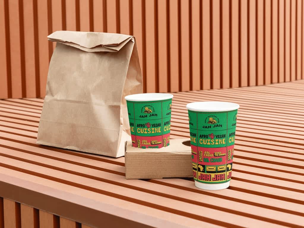 Two green and red branded paper cups on a wooden table next to a brown paper takeout bag.
