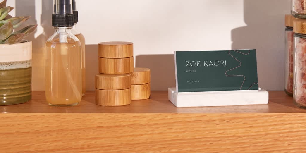Simple dark green business card with thin pink line drawing on wooden counter next to products for a cosmetics business owner.