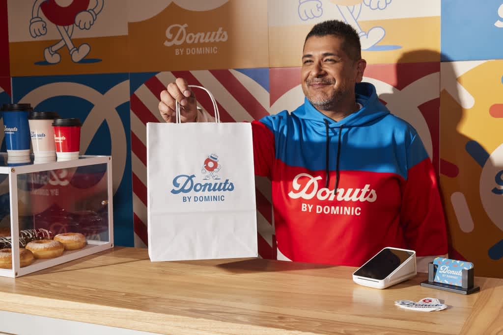 Staff member wearing a blue and red branded hoodie while handing a branded paper bag to a customer.