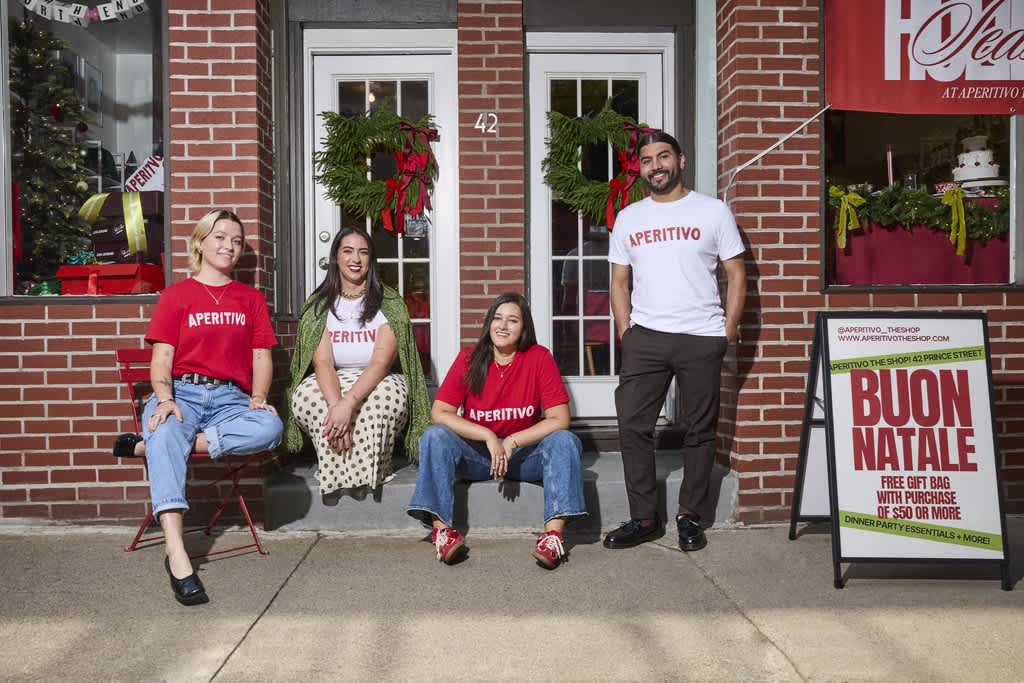 Four people wearing branded T-shirts outside a decorated storefront with holiday signage.