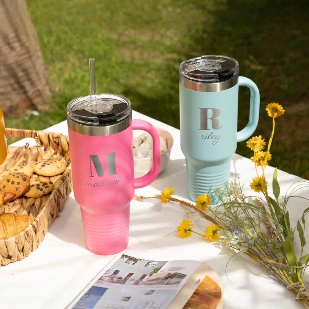 Personalized travel tumblers on an outdoor picnic table next to a basket of cookies and a bunch of flowers. One in pink with silver “Madison” font and other in baby blue with silver “Riley” font.